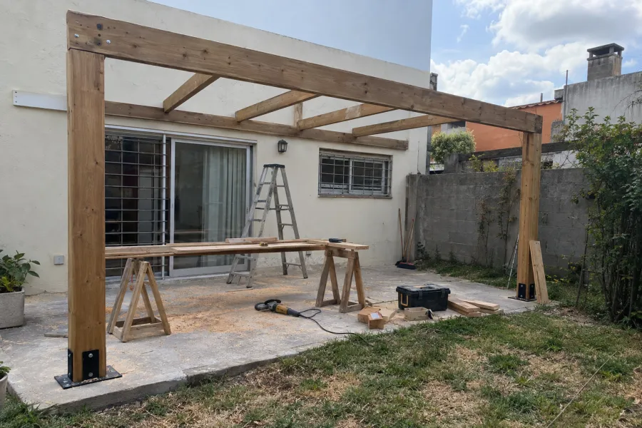 Pérgola de madera en obra: postes en anclajes y vigas colocadas, con escalera, caballetes y herramientas