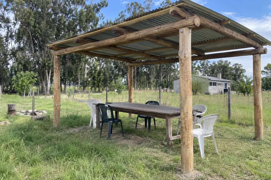 Pérgola de postes rollizos de eucalipto con techo de chapa en un terreno de campo, con mesa larga